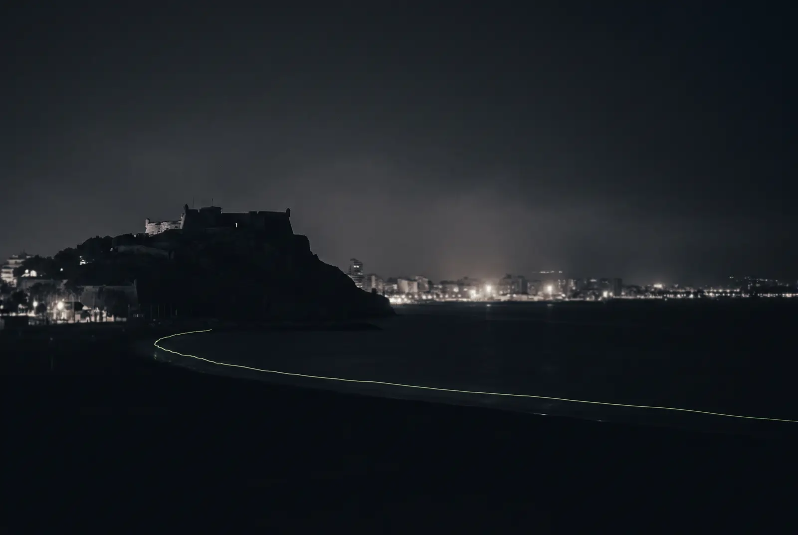 El castillo de Santa Barbara sobre Alicante de noche con una linea lima en la costa